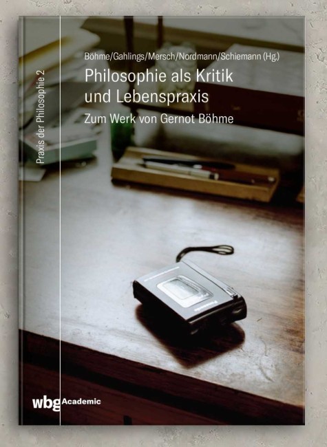 Buchcover "Philosophie als Kritik und Lebenspraxis" - Ein Diktiergerät liegt auf einem Holzschreibtisch, im Hintergrund ist eine längliche Holzschale mit Stiften zu sehen, dahinter steht ein kleines Regal auf dem Tisch, auf dem Glasboden des Regals steht eine Schatulle und ein Gefäß, links neben dem Regal liegt ein Papierstapel und eine kleine Schachtel